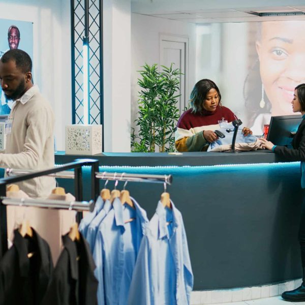 Asian client paying for clothes at store cash register, using credit card at pos terminal in shopping center. Young woman chatting with shop employee, buying modern casual wear.