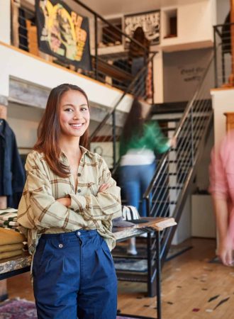 Portrait Of Female Owner Of Fashion Store Standing In Front Of Clothing Display With Customers