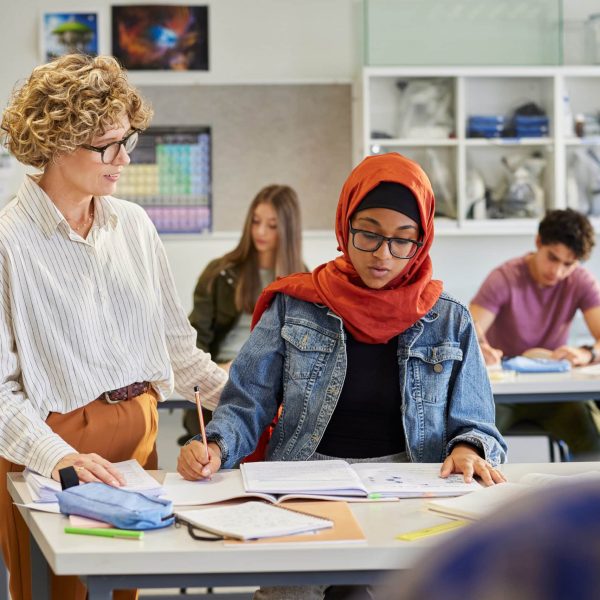 Mature female teacher helping student with her notes in classroom at secondary school. College educator giving support to muslim student in hijab with homework. Mature teacher assisting young learner with studies at high school.