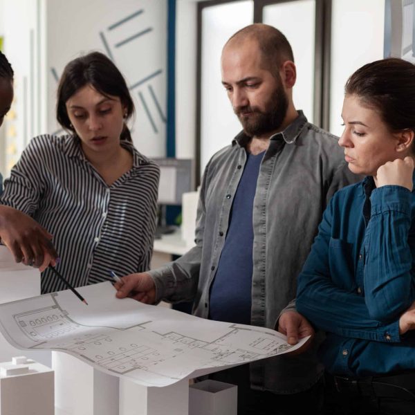 Team of architects analyzing blueprints for building project standing in front of table with real estate maquette. Professional engineers reading construction plans for urban development.