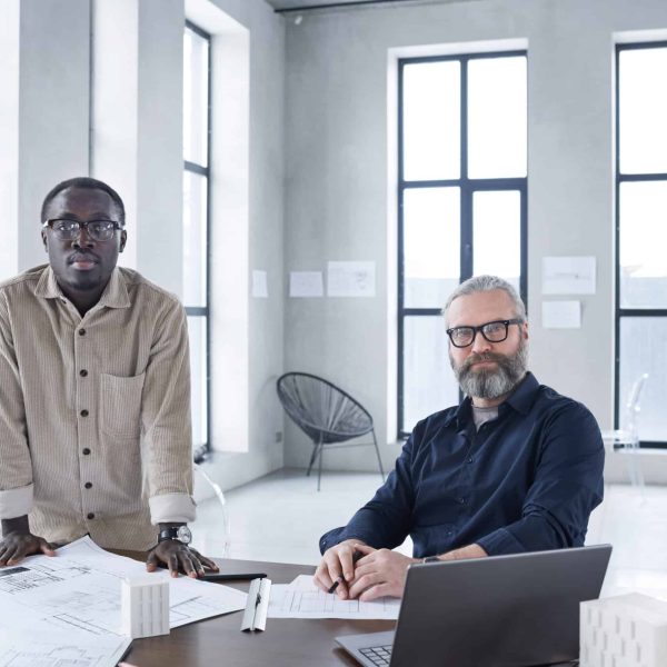 Portrait of two confident businessmen looking at camera while working with blueprints together at office